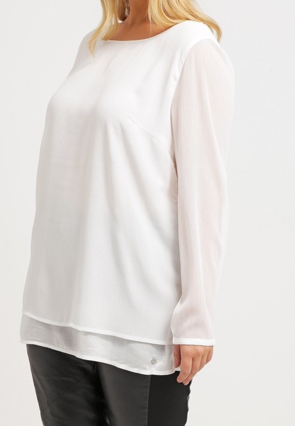 Blusa blanca de manga larga con escote redondeado, que presenta mangas transparentes y un cuerpo liso, combinada con pantalones de cuero negros.