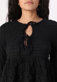 Black textured blouse featuring a keyhole neck with a tie closure, long sleeves, and layered ruffled hem. Soft, crinkled fabric.
