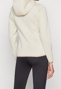 Person in einem strukturierten cremefarbenen Hoodie mit geometrischen Mustern und dunklen, eng anliegenden Hosen, von hinten vor einem schlichten Hintergrund gezeigt.