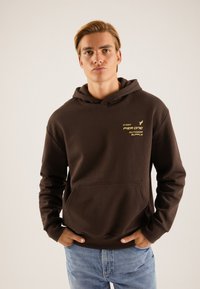 Felpa marrone con tasca frontale, cappuccio e dettagli del logo del marchio giallo. Indossata con jeans azzurri chiari. Tessuto morbido, vestibilità casual.