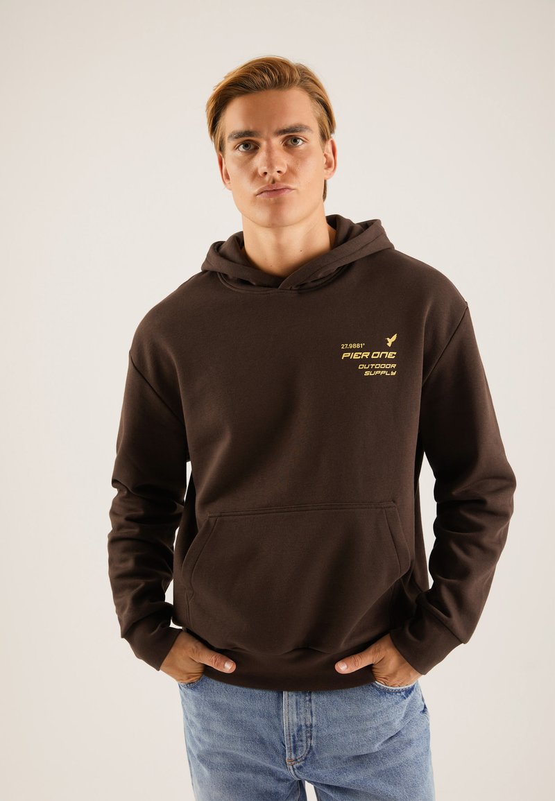 Sudadera marrón con un bolsillo frontal, capucha y detalles del logo de la marca en amarillo. Se lleva con jeans de color azul claro. Tejido suave, corte casual.