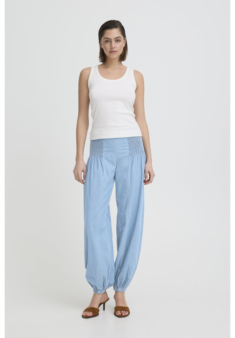 Moteris stovi apsirengusi balta be rankovių palaidine, šviesiai mėlynomis laisvomis kelnėmis su surišamais kulkšnies ryšuliais ir rudu atviru kulnu aukštakulniais sandalais.