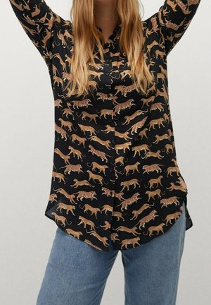 Kvinde iført en sort skjorte med knapper og et print af orange og brune tigre over hele, kombineret med blå jeans, armene hævet.