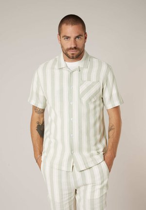 Homme aux cheveux courts et tatouages portant une chemise à manches courtes rayée verticalement vert clair et blanc, assortie d'un pantalon, les mains dans les poches.