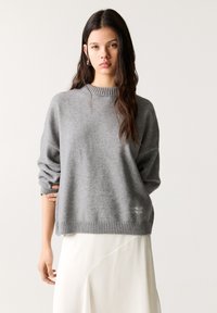 Pulover gri din tricot cu o guler și un tiv ribat, având umeri lăsați și un finisaj texturat. Detaliu tipărit subtil la tiv.