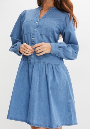 Frau trägt hellblaues Jeanskleid mit langen Ärmeln, Druckknöpfen vorne, zwei Brusttaschen und geraffter Taille.