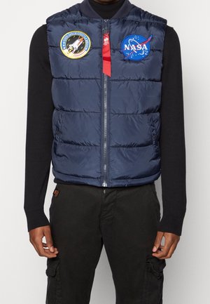 Mężczyzna noszący granatową pikowaną kamizelkę z naszywkami NASA i misji promu kosmicznego na czarnej koszuli z długim rękawem i czarnych spodniach.