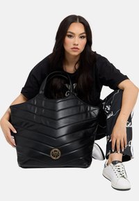 Schwarze gesteppte Handtasche mit Chevron-Muster, aus glänzendem Material gefertigt. Verfügt über ein kreisförmiges goldfarbenes Hardware-Detail an der Vorderseite und robuste Griffe.
