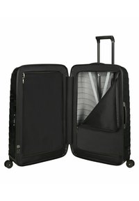 Valise noire à coque dure, avec une surface à motifs. L'intérieur comprend des compartiments zippés, des poches en mesh et des sangles ajustables.