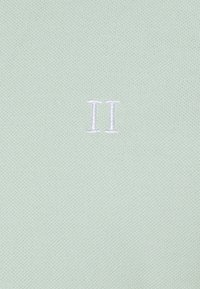 Tissu texturé vert clair avec un motif tricoté. Présente un "II" blanc brodé dans le coin inférieur droit.