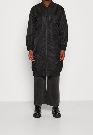 Personne portant une longue veste bomber noire zippée, un pantalon large noir et des bottes noires, debout devant un fond uni.