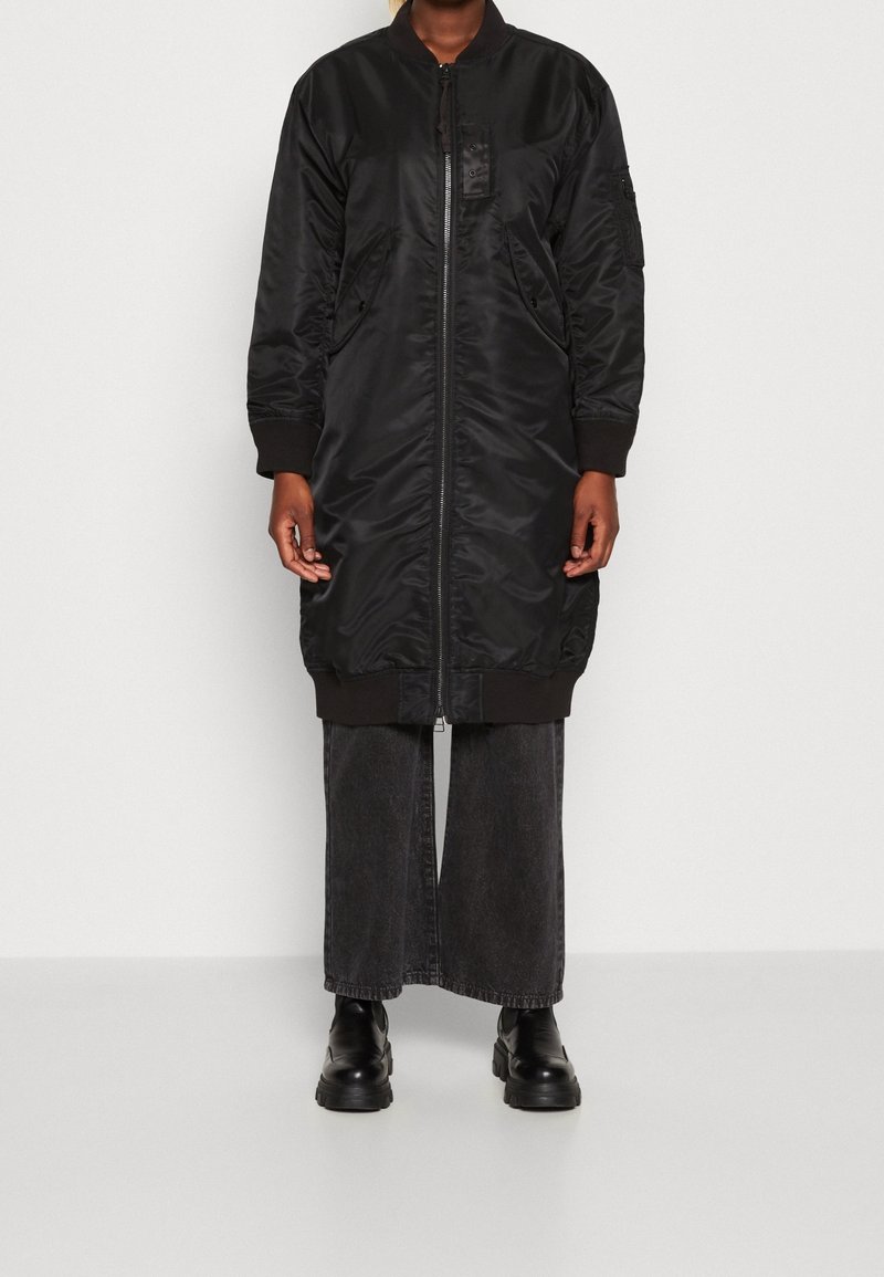 Personne portant une longue veste bomber noire zippée, un pantalon large noir et des bottes noires, debout devant un fond uni.