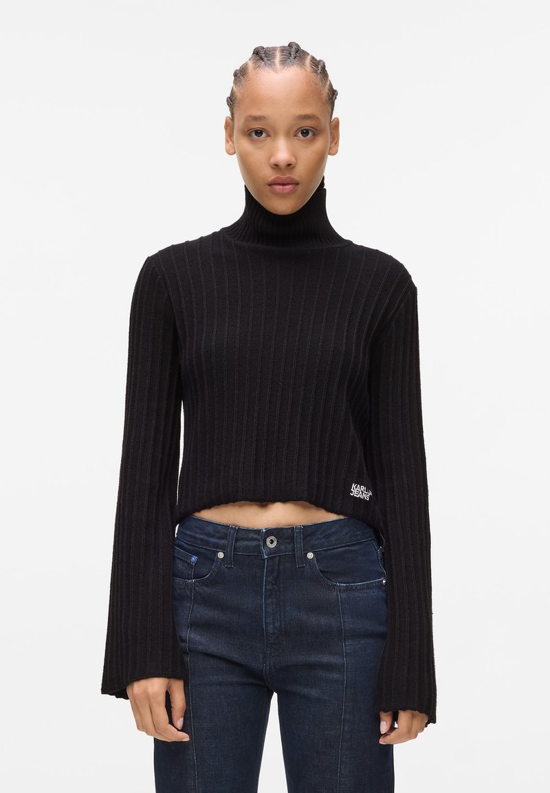 Schwarzer gerippter Rollkragenpullover mit cropped Design und ausgestellten Ärmeln; verfügt über ein Logo-Akzent auf der linken unteren Seite. Kombiniert mit dunklen Jeans.