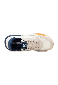 Zapatilla deportiva beige con acentos en azul marino, parte superior texturizada, cordones suaves y suela de goma; presenta un detalle en naranja llamativo cerca de la punta.
