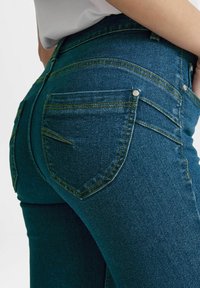 Džínové kalhoty v tmavě modrém odstínu s žlutým prošíváním. Obsahují dvě zadní kapsy a přiléhavý střih. Texturovaná tkanina zvyšuje odolnost.