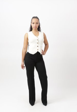 Vrouw met lang krullend haar staat zelfverzekerd en draagt een wit vest met knopen en een zwarte broek met hoge taille tegen een effen witte achtergrond.