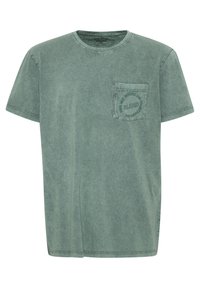 Camiseta verde de manga corta con cuello redondo y un bolsillo en el lado izquierdo del pecho que presenta texto impreso. Material de algodón suave y ligero con un acabado desgastado.