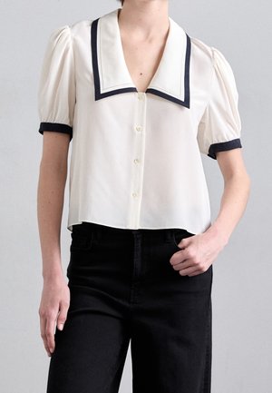 Femme portant un chemisier blanc à manches courtes avec un liseré noir au col et aux poignets, associé à un pantalon noir taille haute.