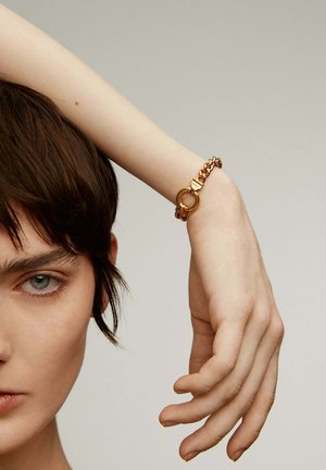 Pulsera de cadena de oro con un cierre circular, llevada en una muñeca delgada. El diseño presenta cadenas entrelazadas y acentos metálicos pulidos.