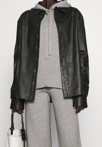 Czarna skórzana kurtka koszulowa, zapinana z przodu, z szarym hoodie pod spód. Model trzyma białą torebkę, prezentując teksturowany materiał.