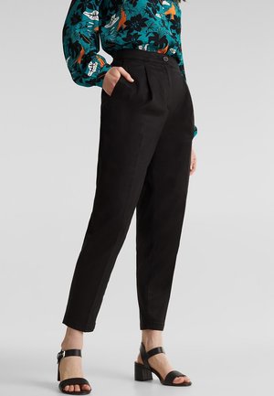 Kobieta nosząca czarne spodnie z wysokim stanem, zwężane, z bocznymi kieszeniami, w połączeniu z czarnymi sandałami na obcasie oraz długim, kwiecistym, turkusowym żakietem.