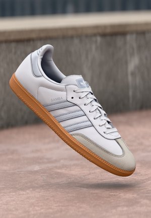 Hvid Adidas Samba sneaker med gummisål og grå accenter svævende over en betonoverflade med sløret baggrund.