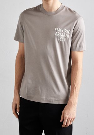 Mężczyzna w taupe krótkim rękawie t-shirtu z białym napisem "EMPORIO ARMANI 1981" na lewej piersi, w połączeniu z czarnymi spodniami.