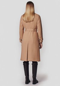 Wełniany płaszcz w kolorze camel z paskiem w talii, zapinany na guziki rękawy i rozcięciem z tyłu. Noszony z czarnymi skórzanymi kozakami do kolan.