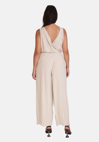 Overall beige con pliegues con un escote pronunciado en la espalda y piernas anchas, hecho de un tejido suave. Los pliegues claros y la textura fluida son prominentes.