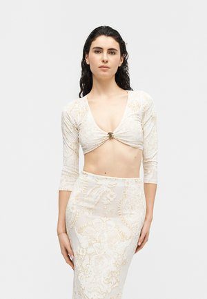 Kobieta o ciemnych falujących włosach, ubrana w kremowy krótki top z rękawami do łokcia oraz pasującą spódnicę z wysokim stanem, ozdobioną złotymi kwiatowymi wzorami.