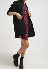 Czarny, oversize kardigan z teksturowanej tkaniny, z dużą kieszenią z przodu. Pod spodem czerwona sukienka w kratę w czarnym i czerwonym kolorze. Czarnie buty dopełniają całość.