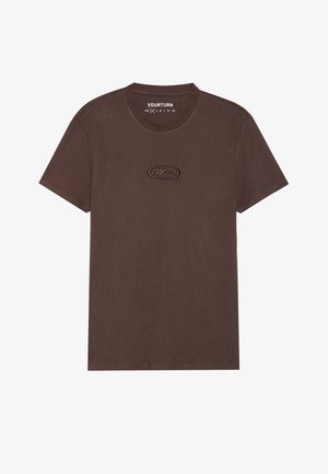 Brun bomulds T-shirt med rund hals, korte ærmer og et tekstureret emblem i midten. Enkelt og afslappet design.