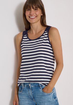 Kvinde smilende iført en navy- og hvidstribet ærmeløs top og blå jeans med den ene hånd i lommen mod en neutral baggrund.