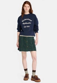 Marinblå sweatshirt med vit text "Timberland Authentic Est 1973", kombinerad med en grön manchesterkjol med knappar framtill.