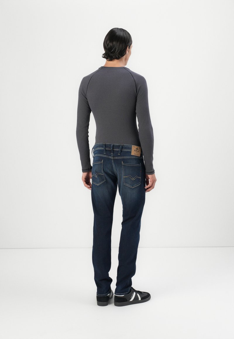 Langärmliges graues Oberteil mit geripptem Muster, kombiniert mit dunkelblauen Jeans, die ein Lederpatch und ein gesticktes Design auf der Rücktasche haben.