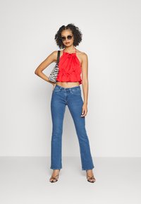 Top cropped halter em seda vermelha com bainha em babados, emparelhado com jeans flare azuis. Os acessórios incluem uma mala listrada em preto e branco e óculos de sol.