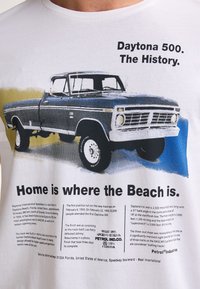 Biała koszulka z grafiką niebieskiego pickupa oraz nadrukiem na temat Daytona 500, zawierająca informacje historyczne i szczegóły dotyczące marki.