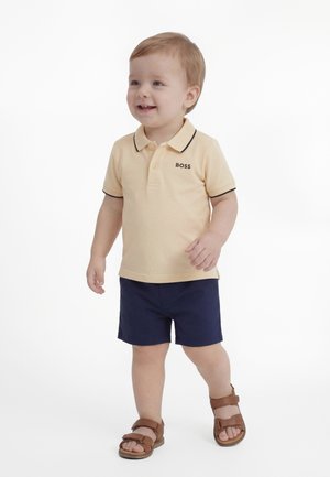 Bambino in piedi, indossa una polo beige con il logo "BOSS", pantaloncini blu navy e sandali marroni, sorride guardando di lato.