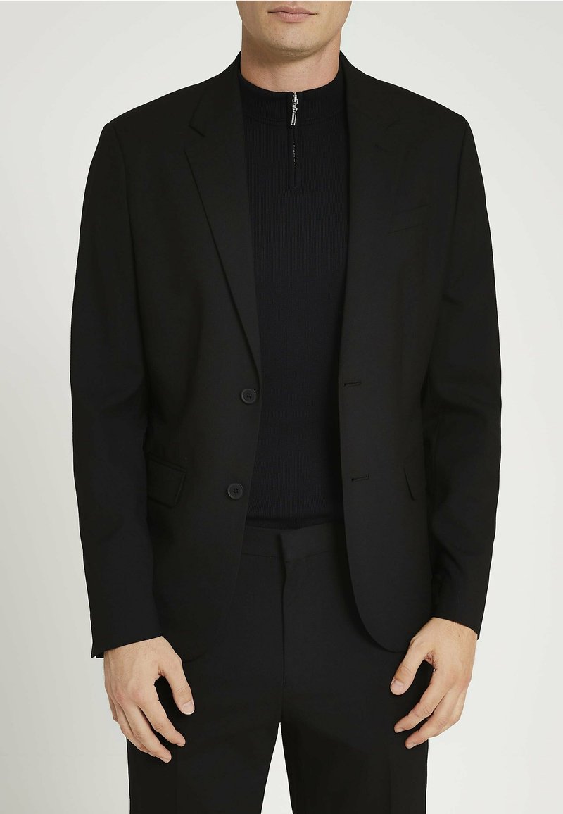 Blazer noir ajusté avec deux boutons, revers en notch et poches latérales. Porté sur un pull à col montant zippé noir. Tissu lisse.