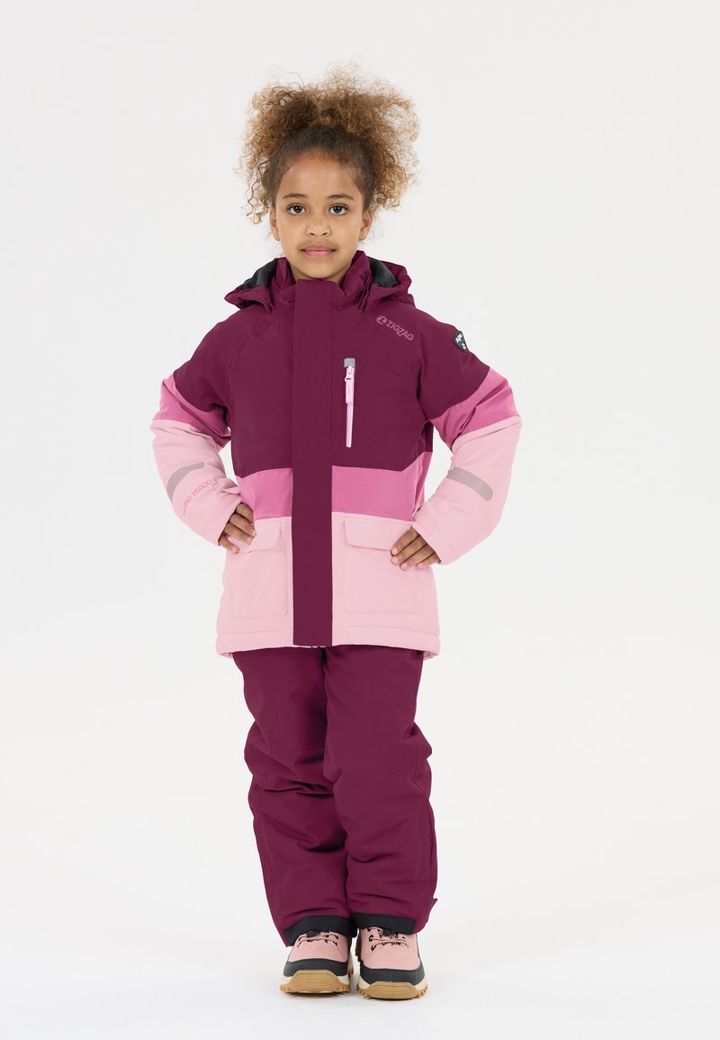 Paarse en roze skijas met capuchon, ritszakje op de borst en roze skibroek. Heeft een stevige textuur en versterkte manchetten.