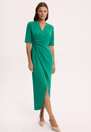 Kobieta stojąca w zielonej sukience kopertowej z rękawami do łokcia i beżowych, szpiczastych butach na obcasie na jednolitym tle.