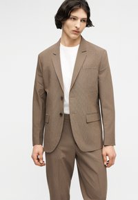 Νεαρός άνδρας που φοράει ένα ανοιχτό καφέ κοστούμι και λευκό πουκάμισο με στρογγυλή λαιμόκοψη, στέκεται με τα χέρια χαλαρά μπροστά σε απλό ανοιχτό φόντο.