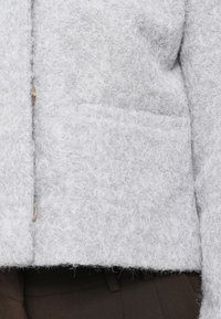 Ανοιχτό γκρι μαλακό μπουφάν με υφή, φτιαγμένο από απαλό υλικό, με μπροστινή τσέπη και κλείσιμο με κουμπί. Μινιμαλιστικός σχεδιασμός με υφή φινιρίσματος.