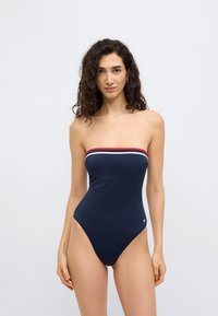 Μπλε σκούρο ολόσωμο μαγιό χωρίς τιράντες, με κόκκινες και λευκές ρίγες στο πάνω μέρος, διαθέτει ψηλή κοπή στα πόδια και απαλό, ελαστικό ύφασμα.