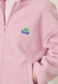 Ružová fleecová bunda s jemnou textúrou, plný zips a bočné vrecká. Obsahuje farebný logopatch na hrudi.
