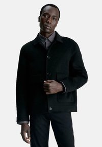 Zwart wollen mantel met knoopsluiting aan de voorkant, twee voorzakken en een kraaglijn. Kenmerkt zich door een ontspannen pasvorm en een gestructureerde stof.