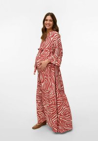 Lang rood en crèmekleurig gekleed met een patroon, met losse mouwen en een strik in de taille, aansluitend over een zichtbare buik, gecombineerd met bruine schoeisel.