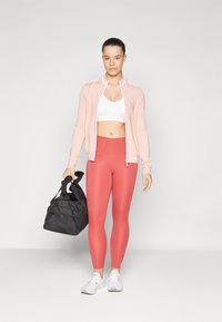 Rózsaszín cipzáros dzseki, fehér sportmelltartó és piros leggings. Fekete edzőtáskát tart. Fehér sportcipők, egyszerű dizájn, testre simuló forma, sima textúra.