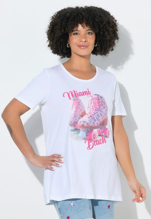 Kvinde med krøllet hår iført en hvid T-shirt med pink rulleskøjter og teksten "Miami Beach", kombineret med lyseblå jeans med stjerne patches.