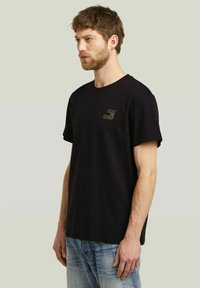 Camiseta negra de algodón, con cuello redondo, mangas cortas y una pequeña etiqueta de tela en el lado izquierdo del pecho. Llevada con pantalones vaqueros azules.